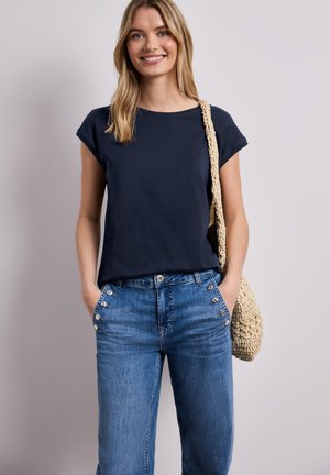 Junge Frau lächelt, trägt ein marineblaues Kurzarmshirt, blaue hochgeschnittene Jeans mit Knopfdetails und trägt eine gewebte Schultertasche.