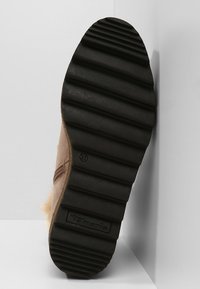 Semelle d'une botte d'hiver marron avec de profondes rainures horizontales et un logo Tamaris, présentée à la verticale sur un fond blanc.
