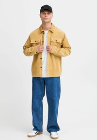 Gele corduroyjas met twee borstzakken, gecombineerd met een witte T-shirt en blauwe wijde jeans. Witte sneakers met groene accenten.
