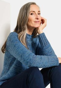 Blauer Strickpullover mit strukturiertem Muster, hohe Ausschnittform und gerippte Bündchen. Dunkle Hose sichtbar, vor einem hellen Hintergrund.