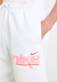 Hvide bomulds sweatpants med elastisk talje, med et stort rødt prikket "nike"-logo og teksten "International Sportswear" på siden.