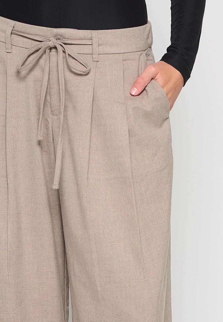 Beige Hochbundhosen mit Falten, einem vorderen Knopf und gebundenem Kordelzug, getragen mit einem schwarzen Langarmoberteil, eine Hand in der Tasche.