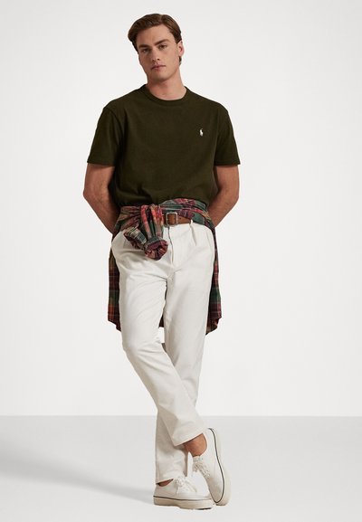 Olivgrön t-shirt, vita byxor, rutigt skjorta knuten runt midjan, brun skärp, vita sneakers; avslappnad outfit med lager av texturer och färger.