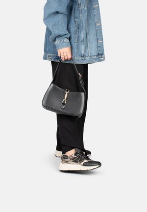 Borsa nera con un design strutturato, hardware dorato e manico superiore, indossata con una giacca di jeans e pantaloni neri, abbinata a sneakers multitessuto.