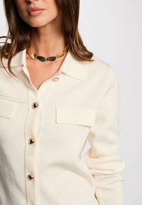 Cardigan lavorato a maglia color crema con collo a camicia, chiusure con bottoni dorati e due tasche anteriori. Completato con una collana dorata chunky.