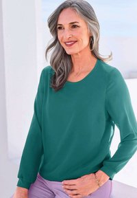 Blusa verde smeraldo con maniche lunghe, scollatura rotonda e texture liscia, abbinata a pantaloni lavanda; include un bracciale delicato.
