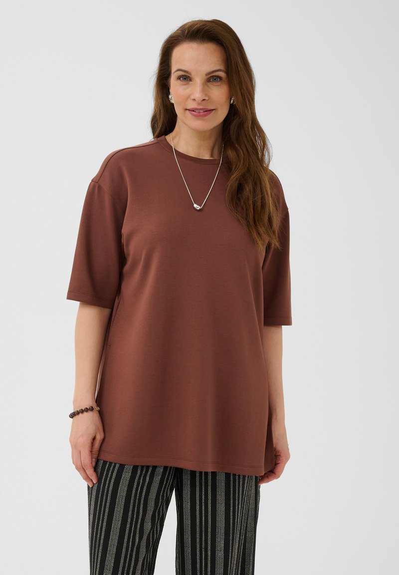Femme aux longs cheveux bruns portant un haut marron ample à manches courtes, un pantalon rayé noir et blanc, un collier en argent et un bracelet, debout devant un fond blanc.