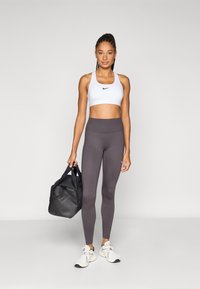 Hvit sports-BH med svart logo, grå høytlivsleggings, hvite joggesko og en svart duffelbag. Ensfarget bakgrunn, modell stående oppreist.