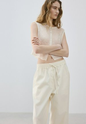 Mujer joven con el cabello largo lleva una blusa sin mangas beige con botones y pantalones holgados de color crema, está de pie con los brazos cruzados mirando hacia un lado.