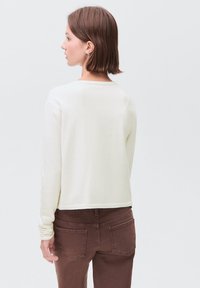 Femeie cu părul scurt și brunet, purtând un top alb cu mânecă lungă și pantaloni maro, stând ușor întoarsă spre stânga pe un fundal simplu.