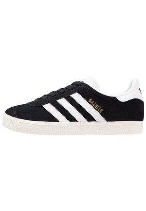 Czarny zamszowy sneaker Adidas Gazelle z białą podeszwą, białą pętelką na pięcie, trzema białymi paskami po bokach i złotym napisem "Gazelle".