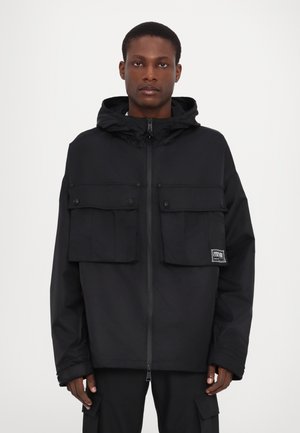 Black hooded jacket made from lightweight material, featuring two large front pockets, a zipper closure, and elastic cuffs.