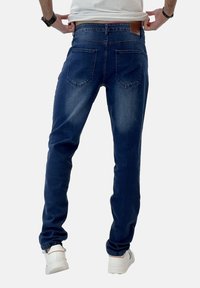 Jeans de denim azul oscuro con corte slim, que cuentan con cinco bolsillos y una etiqueta de cuero marrón en la cintura trasera. Textura suave.