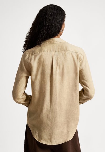 Kobieta o długich, kręconych włosach, ubrana w beżową bluzkę z długim rękawem i ciemne spodnie, widziana od tyłu na jednolitym białym tle.