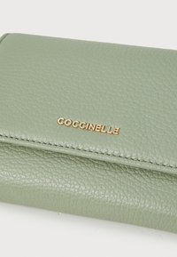 Green textured leather wallet with visible stitching. Gold "Coccinelle" logo prominently displayed on the front. Compact rectangular shape.