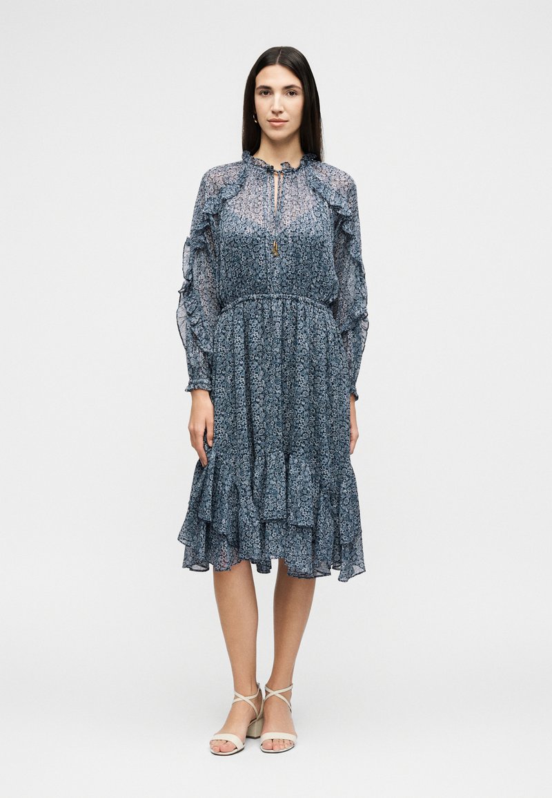 Γυναίκα στέκεται φορώντας ένα μπλε φόρεμα με λουλουδάτο μοτίβο, μακριά μανίκια, βολάν και λευκά ψηλοτάκουνα σανδάλια με λουράκια, ουδέτερο φόντο.