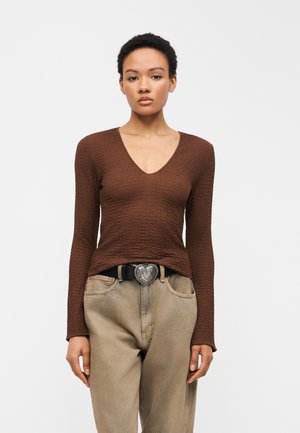 Kvinne med kort krøllete hår som har på seg en tettsittende teksturert brun langermet topp, beige bukser og et svart belte med hjerteformet spenne.