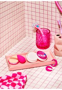 Różowa gumowa gąbka do makijażu i narzędzia na jasnoróżowej tacy. Różowy kieliszek, żyletka i wzorzysta ściereczka na płytkowej powierzchni.