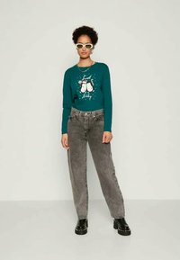 Tirkīza džemperis ar svinīgu dizainu, apvienots ar augsto jostasvietu pelēkajām džinsām. Melni kurpju tipa apavi bez auklām un apaļas baltas saulesbrilles. Neitrāla fons.