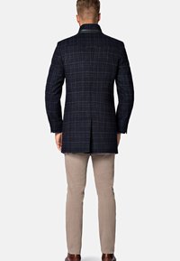 Granatowy płaszcz w kratę z usztywnionym kołnierzem i skórzanymi akcentami, zestawiony z jasnobeżowymi spodniami i brązowymi butami, widziany z tyłu.