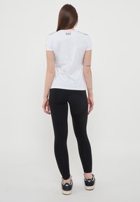 T-shirt bianca a maniche corte con accenti neri sulle spalle e logo EA7; abbinata a leggings neri e sneakers nere con suola bianca.