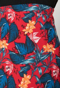 Tela roja con un patrón floral que presenta hojas azules y flores naranjas. Incluye una marca con texto blanco, exhibiendo un diseño vibrante.