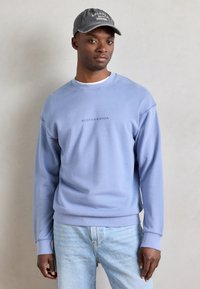 Sudadera azul clara con cuello redondo y mangas largas, con un sutil logo. Combinada con jeans azul claro. Gorra oscura con texto en la cabeza.