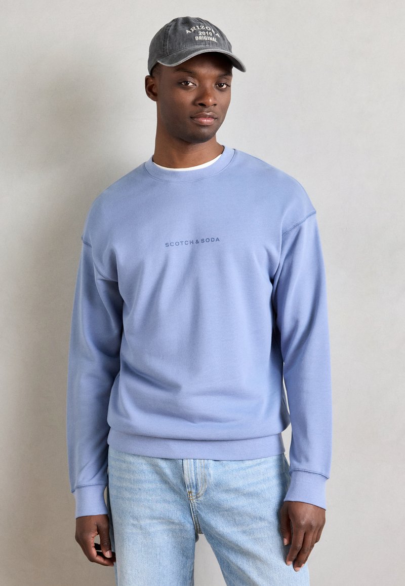 Sudadera azul clara con cuello redondo y mangas largas, con un sutil logo. Combinada con jeans azul claro. Gorra oscura con texto en la cabeza.