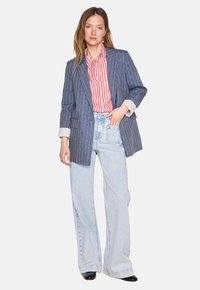 Blazer en denim rayé bleu, avec un design ajusté, des revers à cran et des manches retroussées. Associé à une chemise rayée rouge et un jean bleu clair.