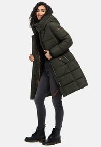 Padded tmavohnedá parka s kapucňou, so žltým zipsom, bočnými vreckami a dlhším strihom. Nosené cez čierny top a šedé džínsy.