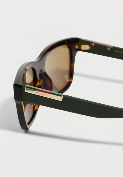 Lunettes de soleil écaille avec des verres marron et des branches vert foncé ornées d'une plaque en or avec le logo Gucci et des rayures rouges et vertes.