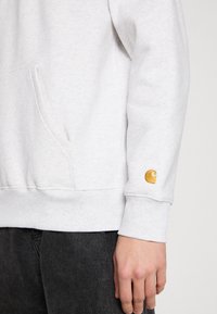 Lys grå sweatshirt med en kængurulomme, ribbede manchetter og et gul logo på ærmet. Blød tekstur og afslappet pasform.