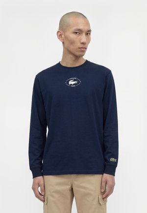 Un mannequin masculin porte une chemise Lacoste bleu marine à manches longues et un pantalon cargo beige sur un fond blanc uni.