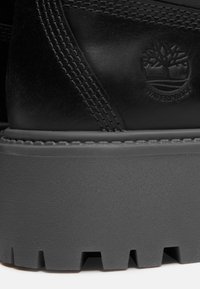 Černá voděodolná kožená bota s texturovanou šedou gumovou podešví, zdobená šitými detaily a vtlačeným logem stromu.