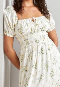 Robe blanche avec imprimé floral jaune, taille smockée ajustée, décolleté carré et manches bouffantes. Tissu léger avec une texture douce.