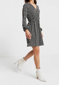 Schwarzes Polka-Dot-Kleid mit langen Ärmeln, tailliertem Schnitt und V-Ausschnitt, kombiniert mit weißen Knöchelstiefeln und einer glatten Textur.
