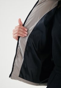 Ľahká bunda na zips s šedým exteriérom a čiernym podšívkou. Interiér má hladkú, lesklú látku, ktorá odhaľuje kontrastné farby.
