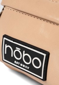 Borsa in ecopelle beige chiaro con un patch logo nero e bianco con il testo "nōbo EST MMXV". Texture liscia, dettagli di cucitura sottili.