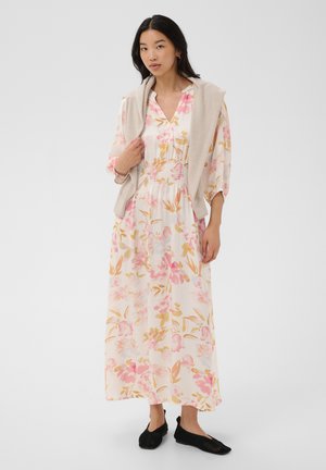 Femme portant une longue robe blanche avec un imprimé floral rose et beige, un pull beige posé sur les épaules, et des chaussures noires sans lacets.