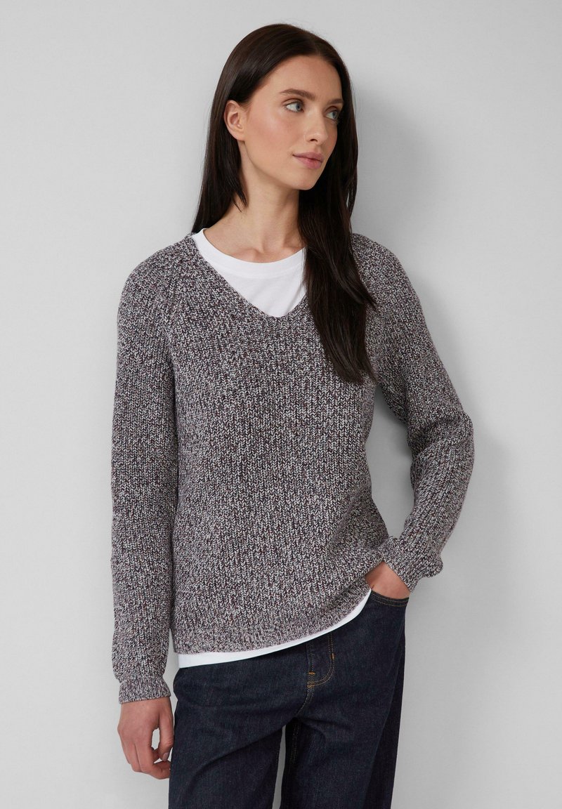 Frau mit langen dunklen Haaren, die einen melierten lila Strickpullover über einem weißen Shirt und dunklen Jeans trägt, steht mit einer Hand in der Tasche.