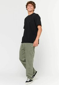 Maglietta nera a maniche corte con un piccolo logo, abbinata a pantaloni in velluto a coste verdi e sneaker nere. Il modello è in una posizione rilassata.