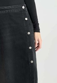 Zwarte denim rok met een sluiting aan de zijkant met zilveren knopen, gestructureerd ontwerp en een gladde textuur. Hand gedeeltelijk zichtbaar.