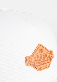 Gorro blanco hecho de tela texturizada con un parche de cuero marrón que presenta un diseño geométrico en relieve y el texto "BLACKSKIES PREMIUM SUPPLIES MMIX."