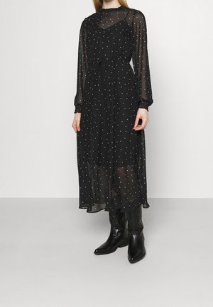 Mujer con un vestido midi negro transparente con lunares blancos, mangas largas y botas de cuero negras hasta media pierna, posando contra un fondo liso.