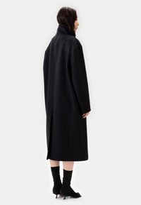 Svart yllekappa med hög krage, lös passform och midjekort längd, med en mittbakssöm och sidospjäll. Svarta strumpor och skor på.
