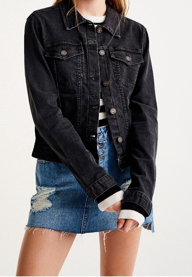 Zwarte spijkerjack met knopen aan de voorkant, twee borstzakken en lange mouwen. Gedragen over een gestreept shirt en gecombineerd met een versleten spijkerrok.