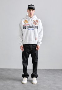 Joven vistiendo un hoodie blanco de DSQUARED2, gorra negra de DSQUARED2, jeans oscuros y zapatillas blancas, de pie sobre un suelo gris contra una pared lisa.