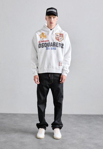 Young man wearing a white DSQUARED2 hoodie, black DSQUARED2 cap, dark jeans, and white sneakers standing on a gray floor against a plain wall.