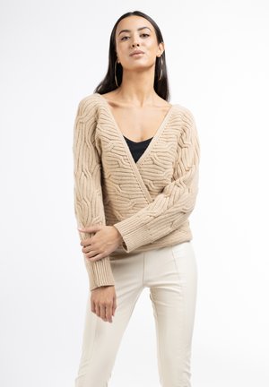 Pulover bej din tricot cu decolteu adânc în formă de V și model texturat de cablu. Mâneci lungi, ușor largi, și manșete cu model zdrobit. Purtați peste un top negru.