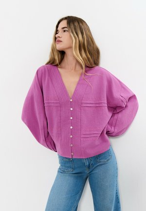 Joven con blusa suelta morada abotonada, de escote en V y mangas globo, que lleva puestos unos vaqueros azules, posando frente a un fondo blanco.